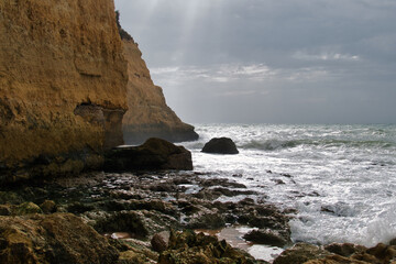 Sunrays reflect on the water of the ocean on a cloudy day. Dark, stony beach and  yellow-brown limestone cliffs
