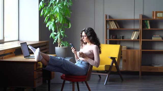 Young female, businesswoman or student relax at workplace drink coffee surf social media sitting at office desk with feet on table. Freelancer writer enjoy free time happy laugh reading text messages