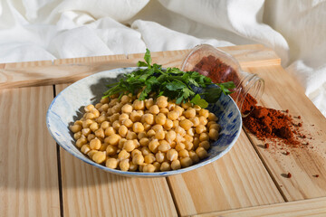 Vertical stills of a plate of boiled chickpeas with parsley fresh leaves and spoiled paprika on a wooden surface. Mediterranean vegan ingredients for cooking hummus cream.
