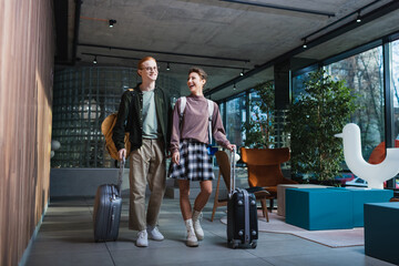 Cheerful woman with backpack and suitcase walking near boyfriend in hotel lobby