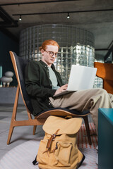Redhead man using laptop near suitcase in hotel lobby