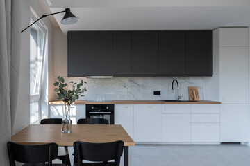 Interior of luxurious kitchen with marble on the wall and dining table with chairs. White furniture and wooden counter in new apartment with window.