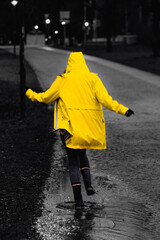 Fotografie von einer Frau, welche an einem regnerischen Tag durch die Pfützen in einem gelben, leuchtenden Regenmantel springt.