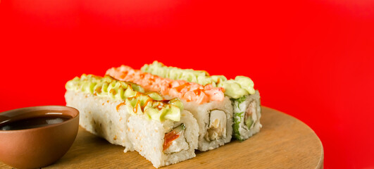 Sushi rolls near soy sauce. Set of tasty sushi rolls with cheese topped with sauce served on wooden table against red background in light room