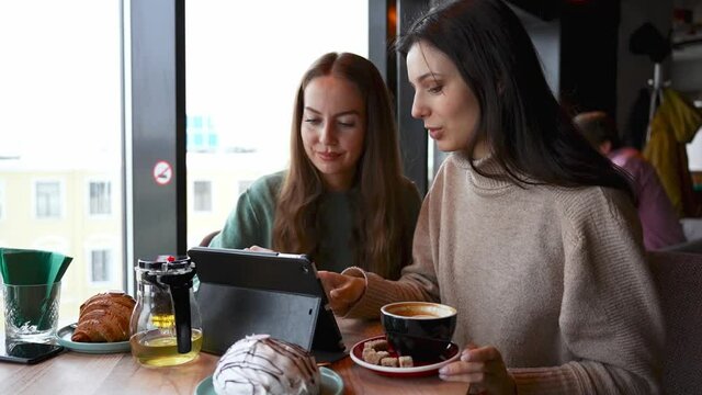 Two young beautiful girls in a cafe are looking at a tablet. One explains to the other how to use the app. The concept of education, co-education in digital technologies