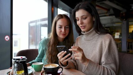 Two young attractive girls in a cafe are looking at the phone. One tells the other, teaches how to use, explains. - Powered by Adobe