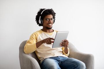 Young Black Guy In Wireless Headphones Relaxing With Digital Tablet In Armchair