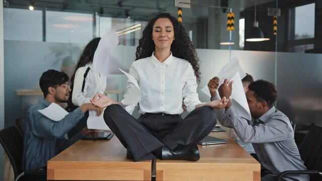 Young hispanic woman sitting on table in lotus position female leader meditating in office businesswoman feels calmness and serenity annoyed angry colleagues throw documents patience workplace concept