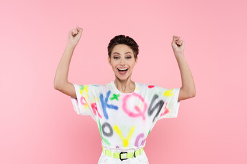 excited young woman with short hair showing triumph gesture while looking at camera isolated on pink