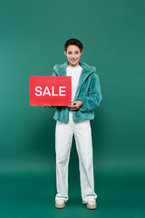 full length view of happy woman in white trousers and faux fur jacket holding red card with sale lettering on green