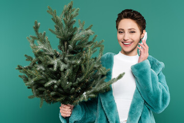 fashionable woman with small fir tree smiling while talking on mobile phone isolated on green