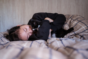 Young Asian woman playing with young black and white cat on top of bed with warm sheets in bedroom