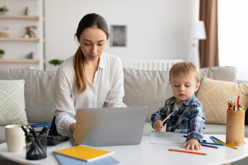 Working mother concept. Young lady typing on laptop keyboard while her toddler son drawing with pencils