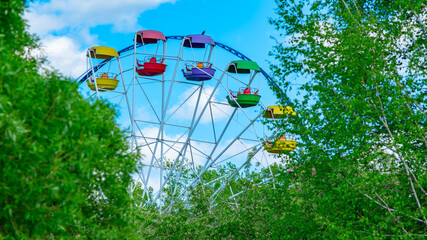 view of metal, old, open colored Ferris wheel booths of the city park against the background of fresh spring green foliage of trees, blue sky with cumulus clouds and bird, crow, magpie sitting on top - Powered by Adobe