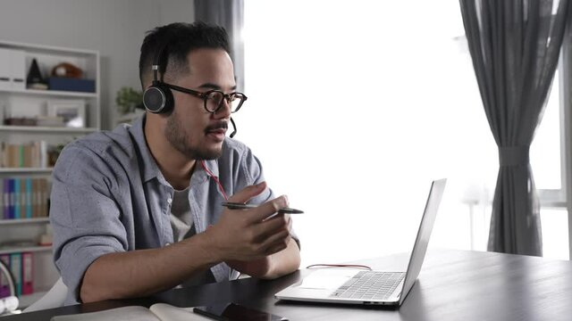 Feeling satisfied and positive at work. Young Asian businessman wearing headset on video call with clients on laptop. Young Asian man in glasses giving online educational class lecture