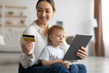 Happy mother with child using digital tablet and credit card, purchasing in internet, sitting on floor in living room