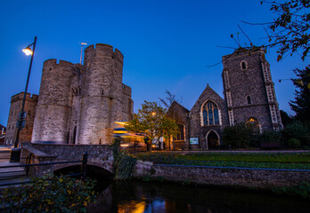 A bus passing by Westgate Towers in Canterbury, with the Great Stour river in the foreground. Night.