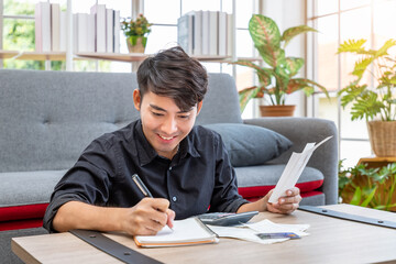 Asian man holding bill and calculating credit card expenses receipt using calculator sitting in living room at home, personal finance concept