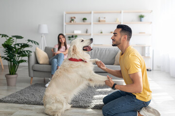 Happy young Arab guy playing with dog, having fun on floor at home, his girlfriend drinking coffee...