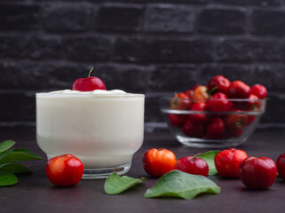 Yogurt in a glass topping with ripe red acerola cherry and cherries, green leaves scattered on a table with black brick background
