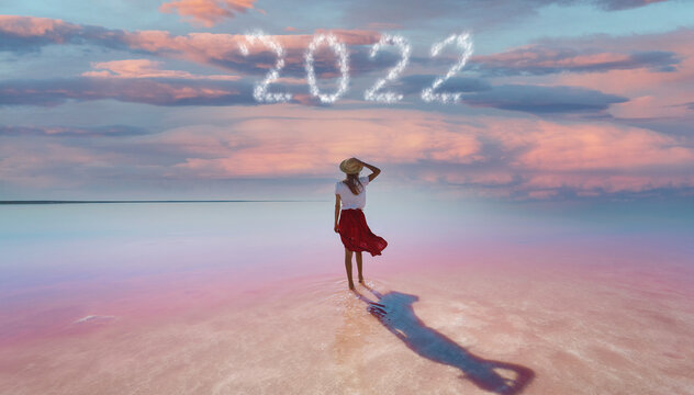 Woman traveler standing in beautiful pink lake with evening purple violet sunset sky with happy new year 2022 numbers