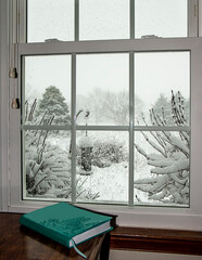 View through a window after a snowstorm and a teal-blue book sitting on the desk in the forefront.