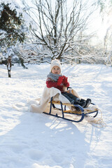 Fototapeta na wymiar Toddler boy sitting on the sleigh in a snowy city park during sunny winter day