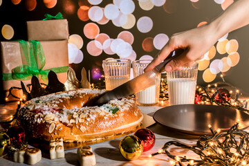The "Rosc&oacute;n de reyes" is a traditional Spanish Epiphany cake with decorative candied fruits. Hand cutting a piece of "rosc&oacute;n de reyes". The Spanish concept of Three Kings Day. Three glasses of milk.