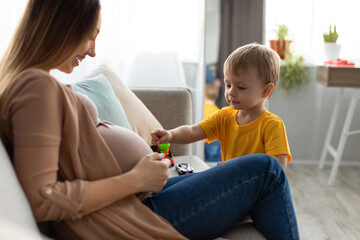 Pregnant mother and her toddler son spending time together at home, little boy touching gently...