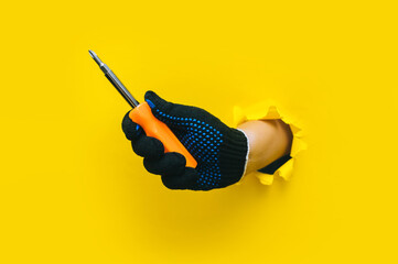 A right hand in a black cloth knitted glove holds a orange screwdriver close up. Torn hole in...