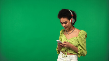 curly african american woman in blouse and wireless headphones using smartphone isolated on green