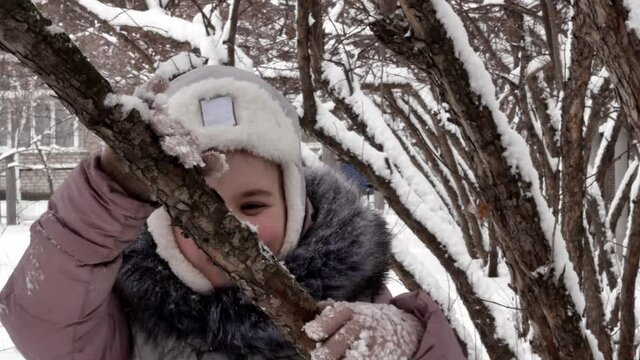 A beautiful happy little girl in winter in warm clothes, a down jacket and a hat with earflaps is having fun among the trees, hugging a branch. Street walk child happy childhood.