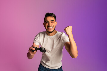 Excited millennial Arab man with joystick celebrating video game win, gesturing YES in neon light