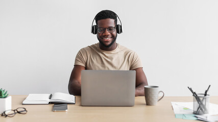 Smiling young black male in headphones communicating online on laptop at desk against white studio background