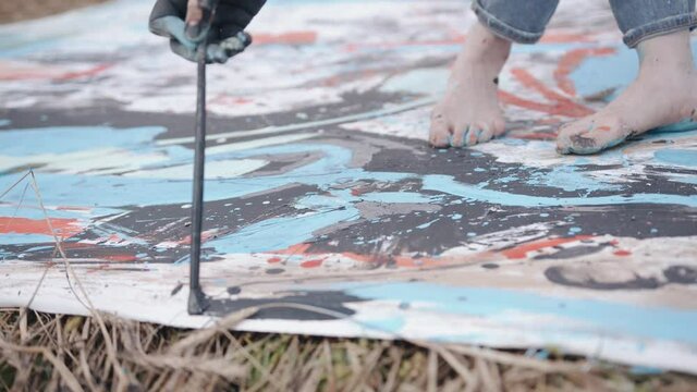 A talented young artist with bare feet stained with paint draws a creative abstract picture with a brush on a canvas in a wheat field