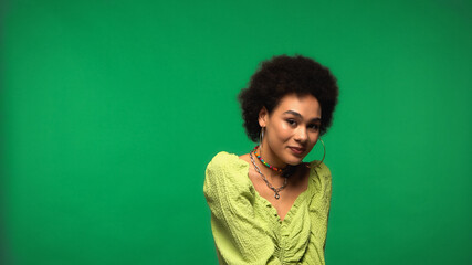 Fototapeta premium positive african american woman in blouse and hoop earrings smiling isolated on green