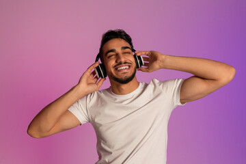 Peaceful young Arab guy listening to music in wireless headset, closing eyes and relaxing in neon light