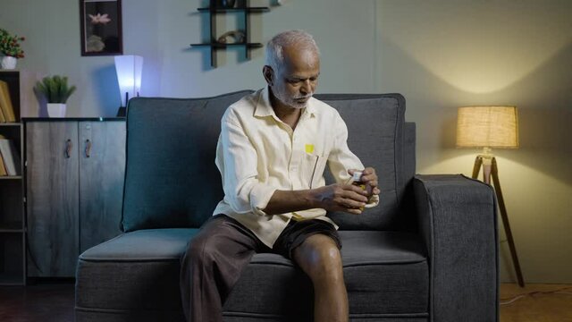 Old man using ayurvedic oil massage for knee joint paint at home on sofa - concept of using natural pain relief oil, osteoarthritis treatment and healthcare