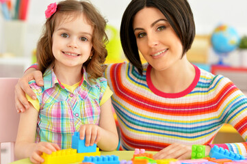 Fototapeta premium Close up portrait of cute little girl and her mother playing colorful plastic blocks together in her room