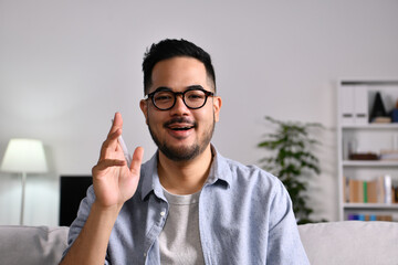 Portrait shot of happy friendly and confident Asian businessman sitting on a couch at home, looking and talking at camera and waving hand saying hello during video call - 478529065