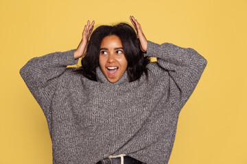 Excited young pretty dark skinned girl in warm knitted sweater isolated on yellow background.