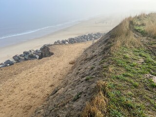 Cliff top view of vast sandy beach landscape with rocks and mist haze over shore and ocean on cold damp foggy weather in Winter at Happisburgh Norfolk East Anglia uk with blue light sky in morning