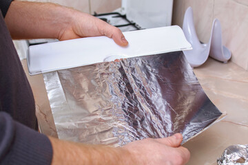 A man tears off the foil from a plastic dispenser to wrap potatoes in it and bake them in the oven.