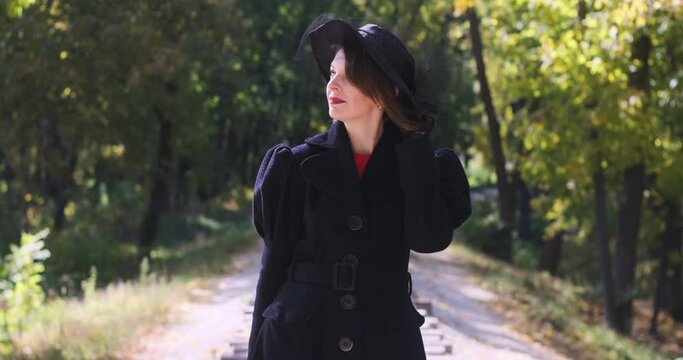Middle-aged woman in black coat stands on forest background and holds hat flying off from the wind. Video in retro style