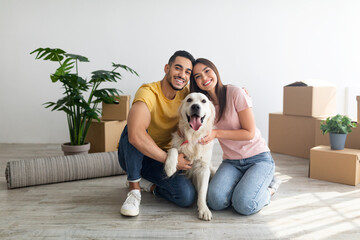 Cheery young international couple with cute golden retriever dog sitting on floor of new home on relocation day