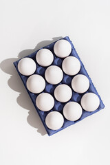 Una docena de huevos blancos de gallina en una huevera de cartón azul sobre una mesa blanca. Vista...