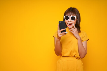 Young beautiful asian woman using smartphone to shopping online, get discount sale and shopaholic or news on a yellow background with surprised and shocked face, OMG