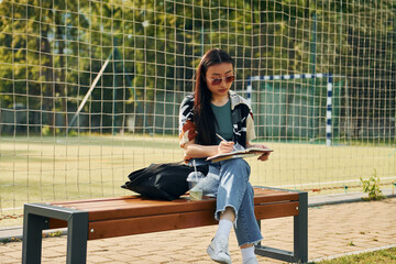 Writes in the notepad. Young asian woman is outdoors at daytime