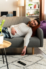 Bearded man sleeping on sofa with tv remote control at hands at home during the day