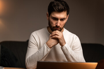 Young male is using laptop in evening at home and looking at digital screen with tired emotions